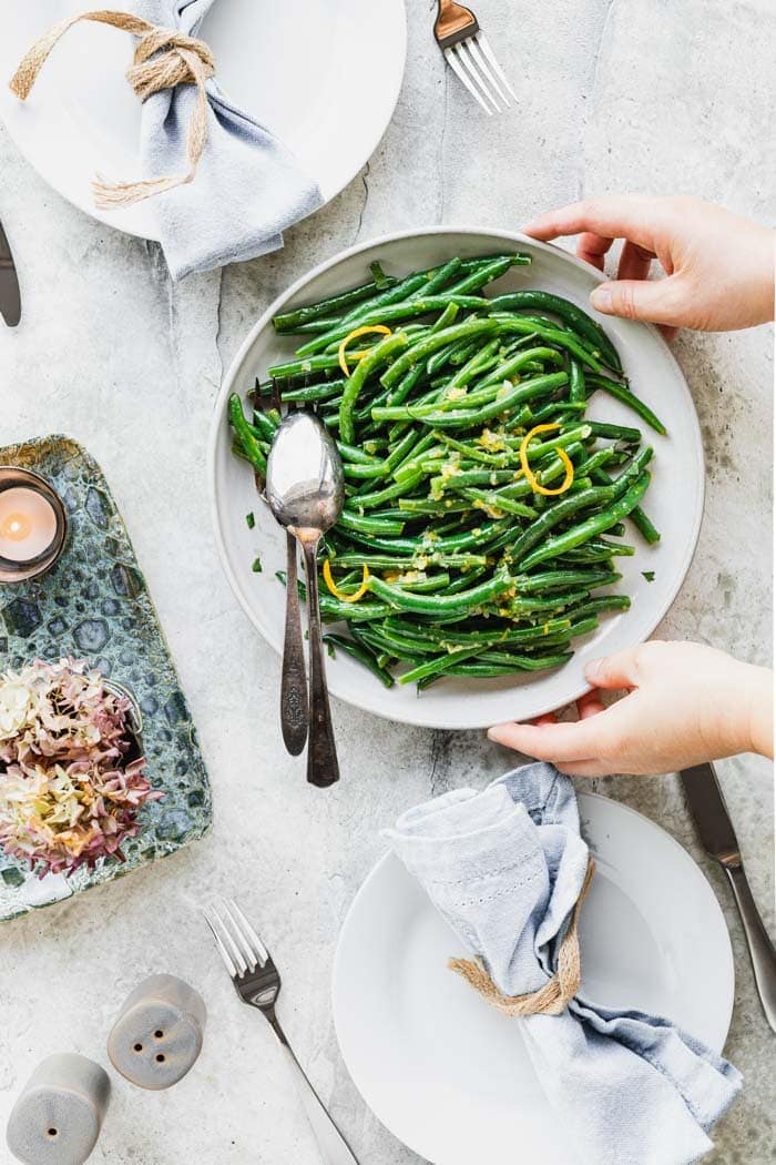 a table with two place settings with blue napkins and two hands are setting down a platter of green beans