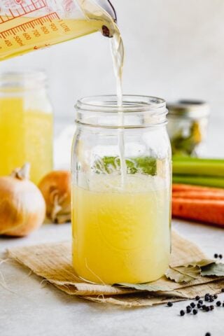 Pouring chicken stock into a jar with other ingredients visible in the background