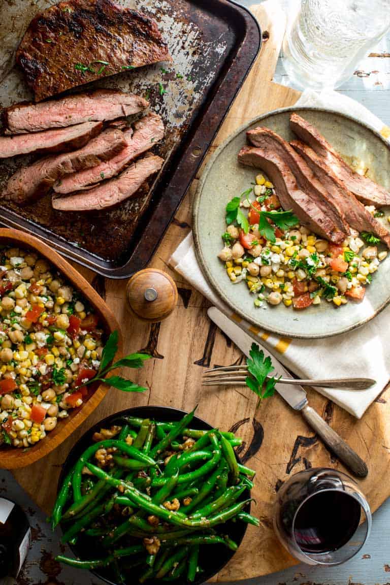 a table scape with a tray of grilled steak, green beans, a bowl of corn salad and a plate with both from overhead