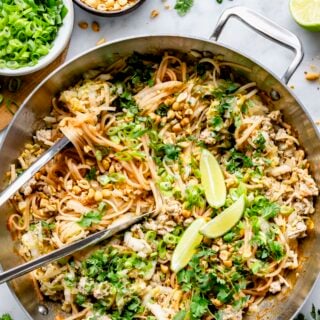 pad thai in a skillet with lime wedges, fresh herbs and tongs
