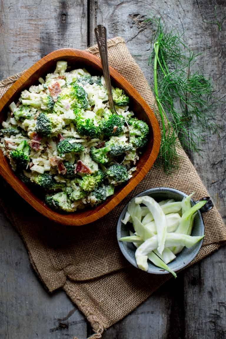 An overhead view of a wooden bowl with broccoli salad and a blue ceramic dish with sliced fennel in it on a gray wooden table