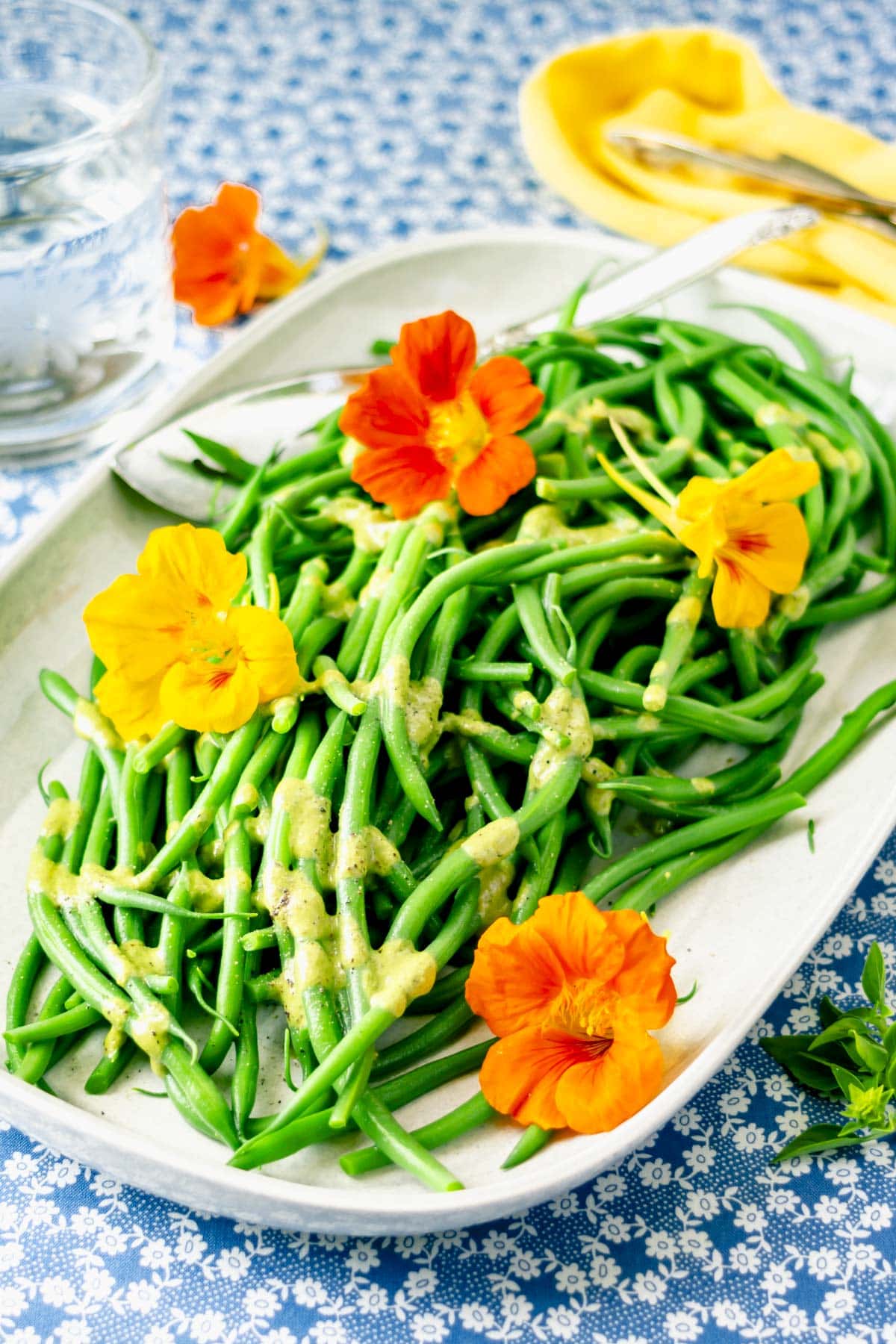 a light gray oblong platter with green beans topped with dressing and edible flowers on a white and blue table cloth and yellow napkin