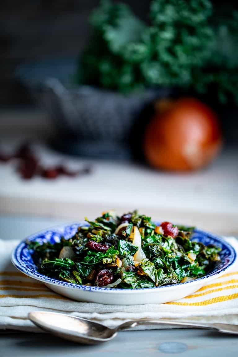 Side view of a bowl of kale with cranberries and balsamic vinegar on a gray table with a gray background