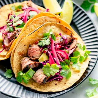 the steak tacos on a black plate with lime wedges and pickled onions