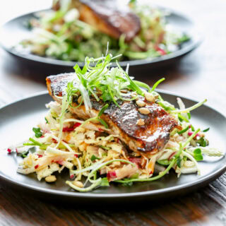 the blackened salmon on top of the shredded slaw with scallion frizzle on top on a black plate on a wooden tabletop