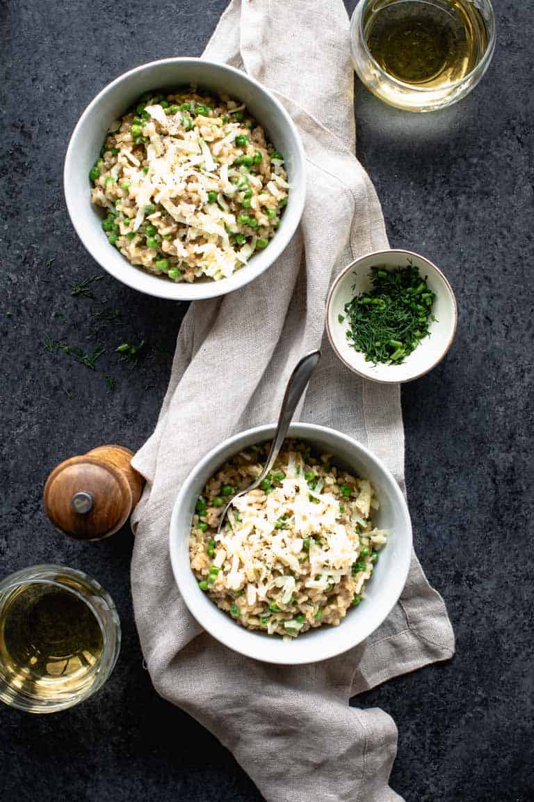 An overhead view of two bowls of brown rice risotto on a black countertop with a tan linen napkin and two glasses of white wine