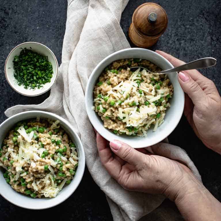 An overhead view of two bowls of brown rice risotto on a black countertop with a tan linen napkin and two hands grabbing a bowl