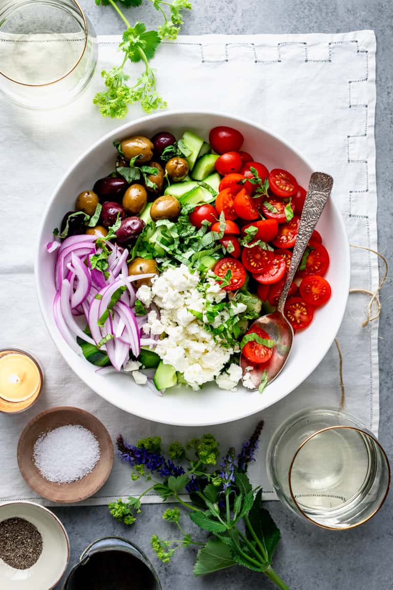 an overhead tablescape with a bowl of Greek salad, flower blossoms, wine and a dish of salt