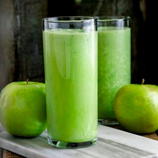two green smoothies with green apples on a white marble cutting board with a dark background