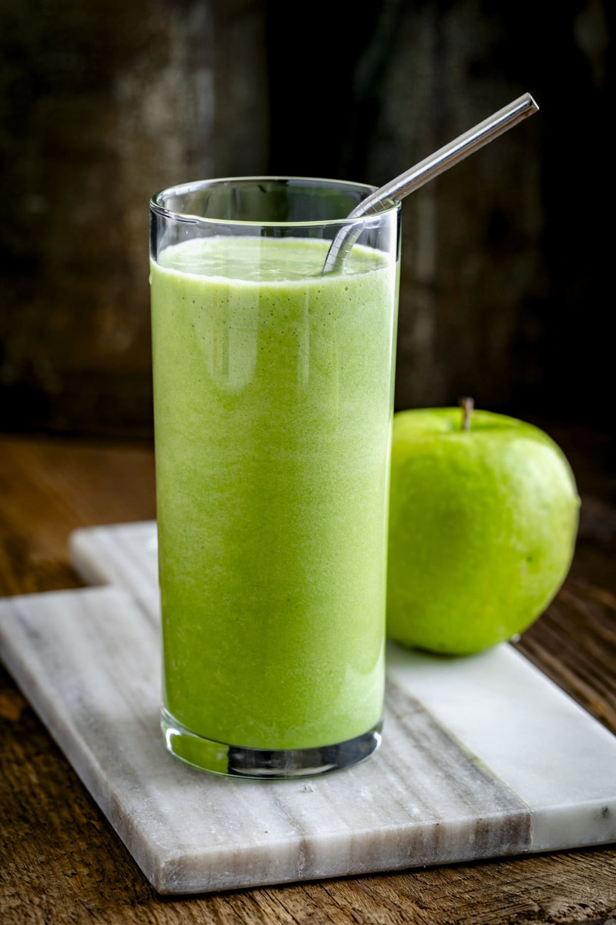 the smoothie in a glass with a metal straw and a green apple on the board behind it