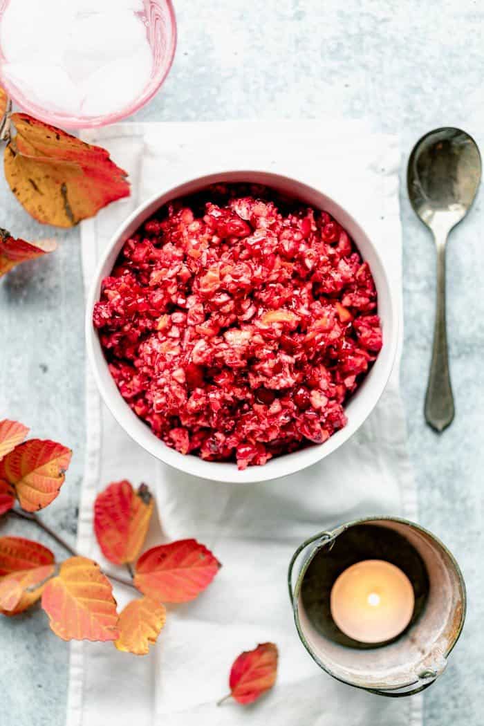 Raw cranberry relish in a white bowl on a blue gray table with orange foliage and a votive candle