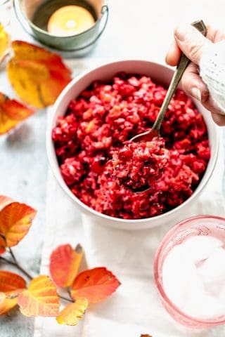 a table with orange foliage and a bowl of raw cranberry relish with a hand holding a spoon of relish