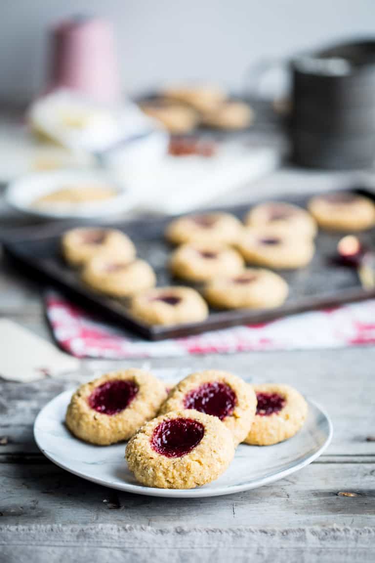 Jam Thumbprint Cookies on a platter with baking ingredients and cookies on a dark baking sheet in the background