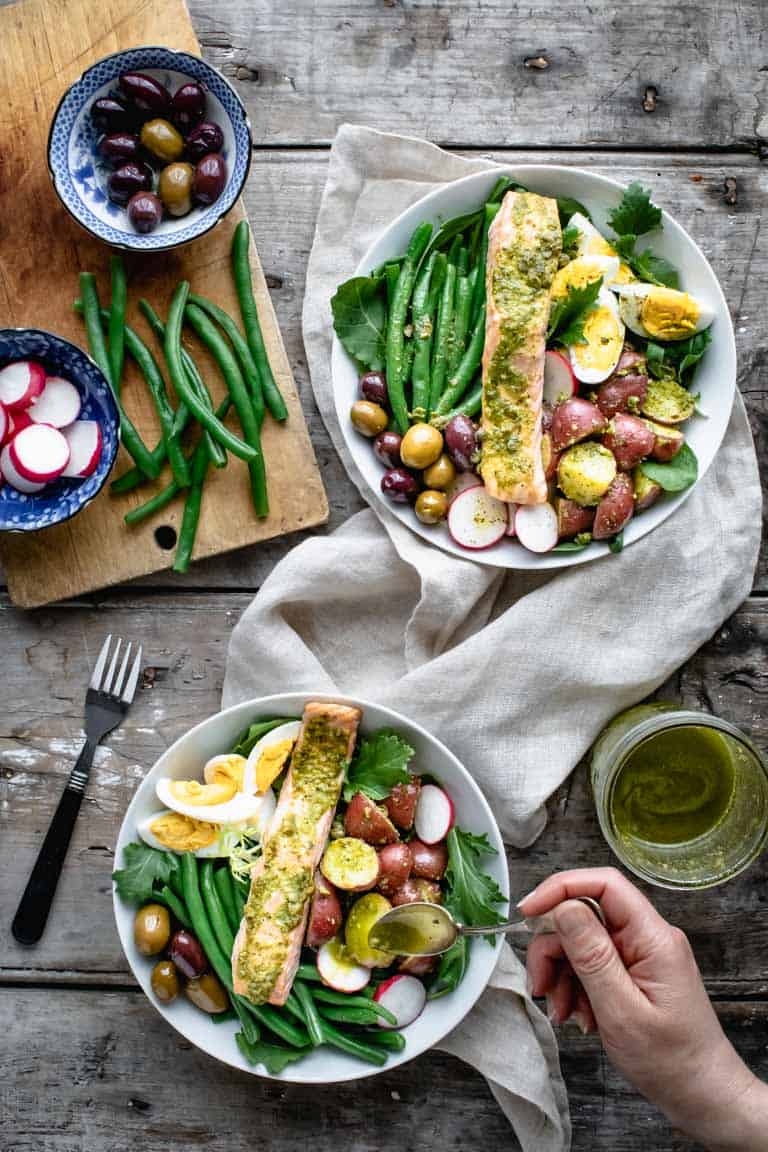 Overhead tablescape with two bowls of Salmon Nicoise being dressed with salad dressing