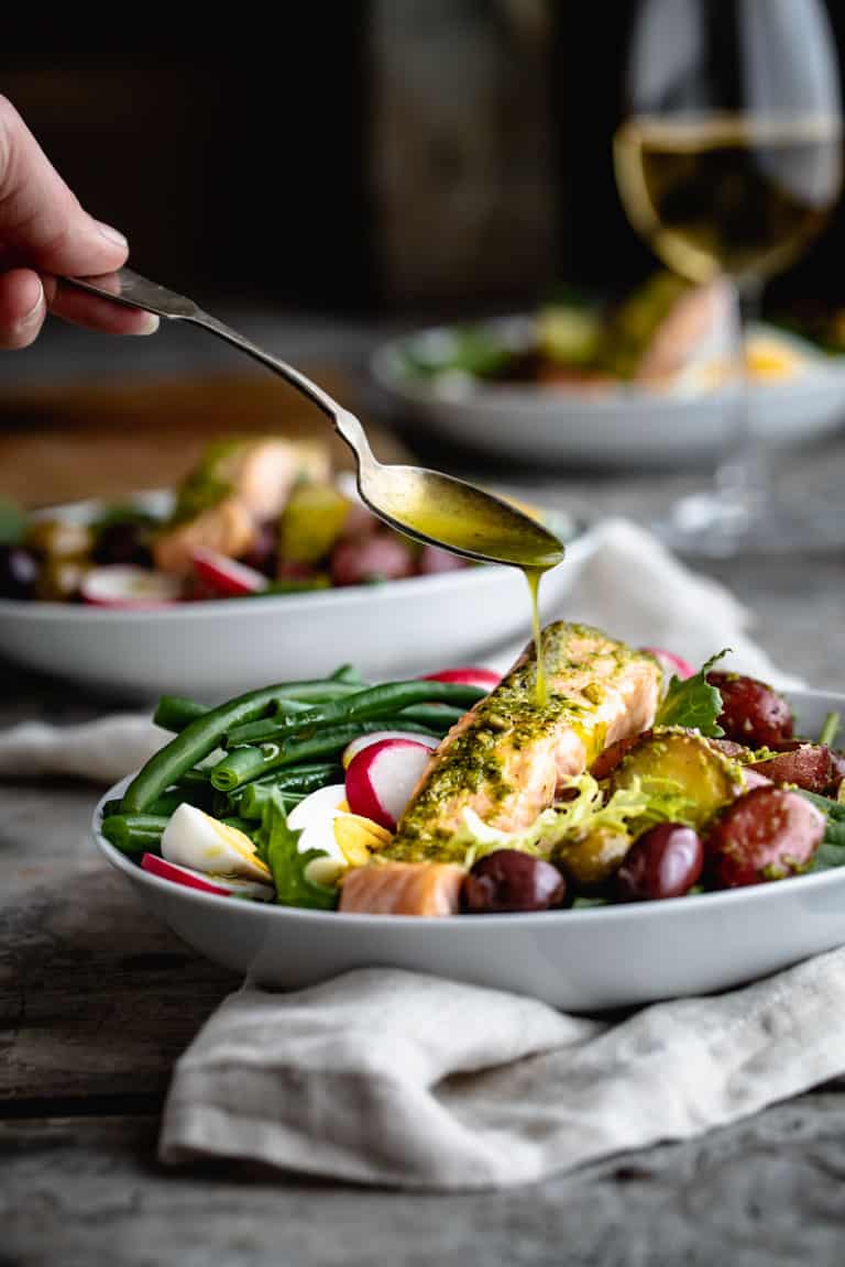 Drizzling dressing over the Salmon Nicoise Salad topped with hard boiled eggs, green beans, olives, potatoes and radishes.