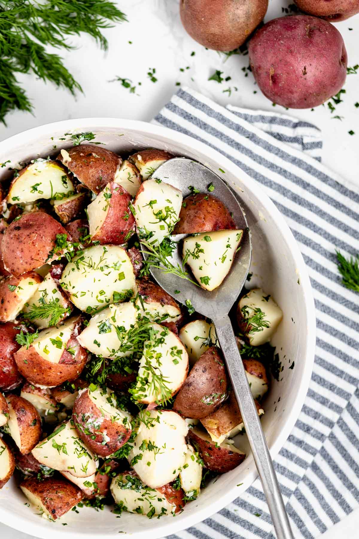 Steamed potatoes in circular white serving bowl with a spoonful of potatoes in serving spoon.