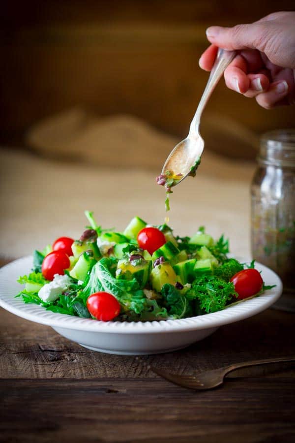 Hand drizzling olive salad dressing from spoon onto a salad in a bowl