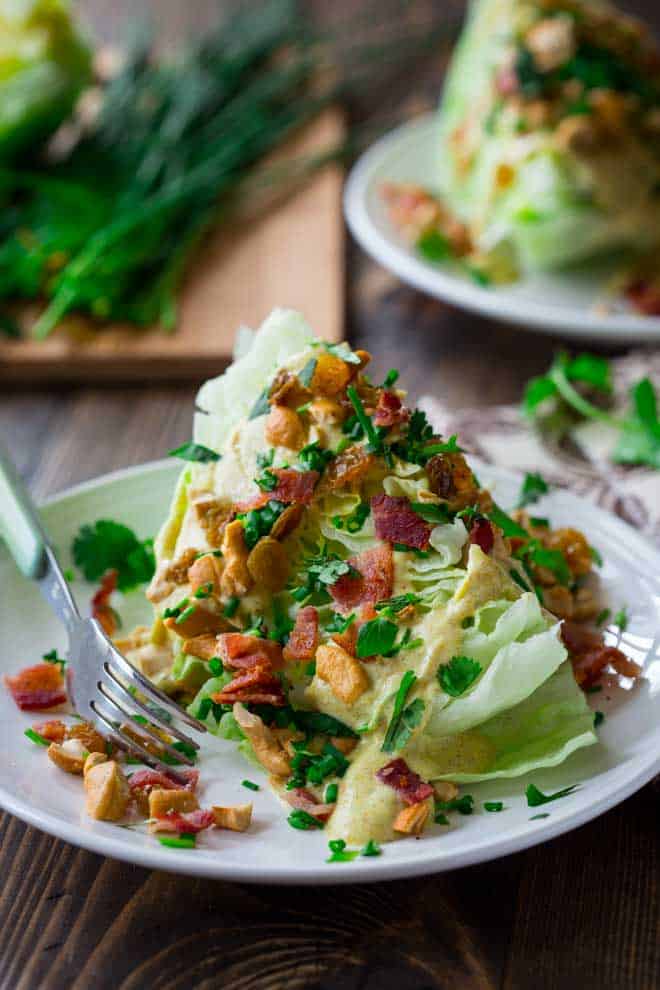 curry wedge salad on a white plate with bacon, cashews, chives, curry dressing, and cilantro on top and a fork to the side