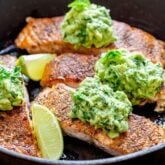 close up of the salmon in the cast iron skillet with the avocado mash on top and lime wedges