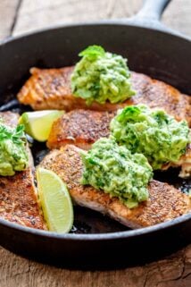 close up of the salmon in the cast iron skillet with the avocado mash on top and lime wedges