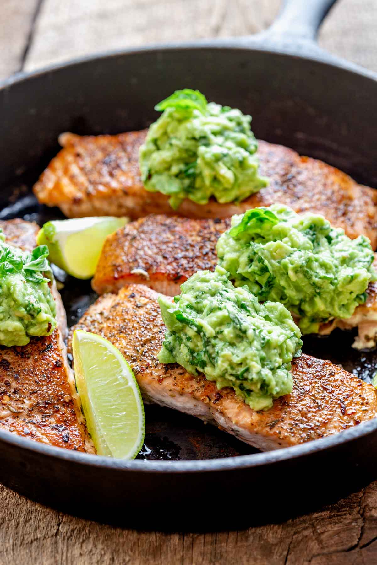close up of the salmon in the cast iron skillet with the avocado mash on top and lime wedges