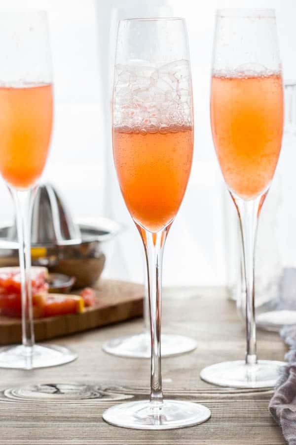 Three champagne flutes filled with Grapefruit Champagne Cocktail with a bottle of
Campari and a citrus juicer in the background