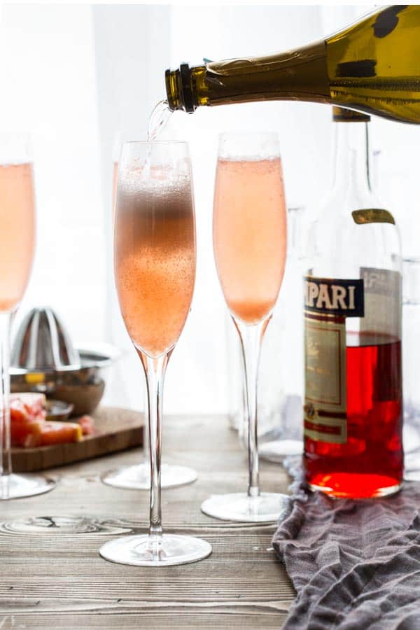 Three champagne flutes filled with Grapefruit Champagne Cocktail and champagne being poured into one with a bottle of
Campari and a citrus juicer in the background