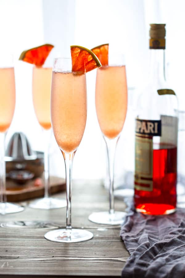 Three champagne flutes filled with Grapefruit Champagne Cocktail with a bottle of Campari and a citrus juicer in the background
