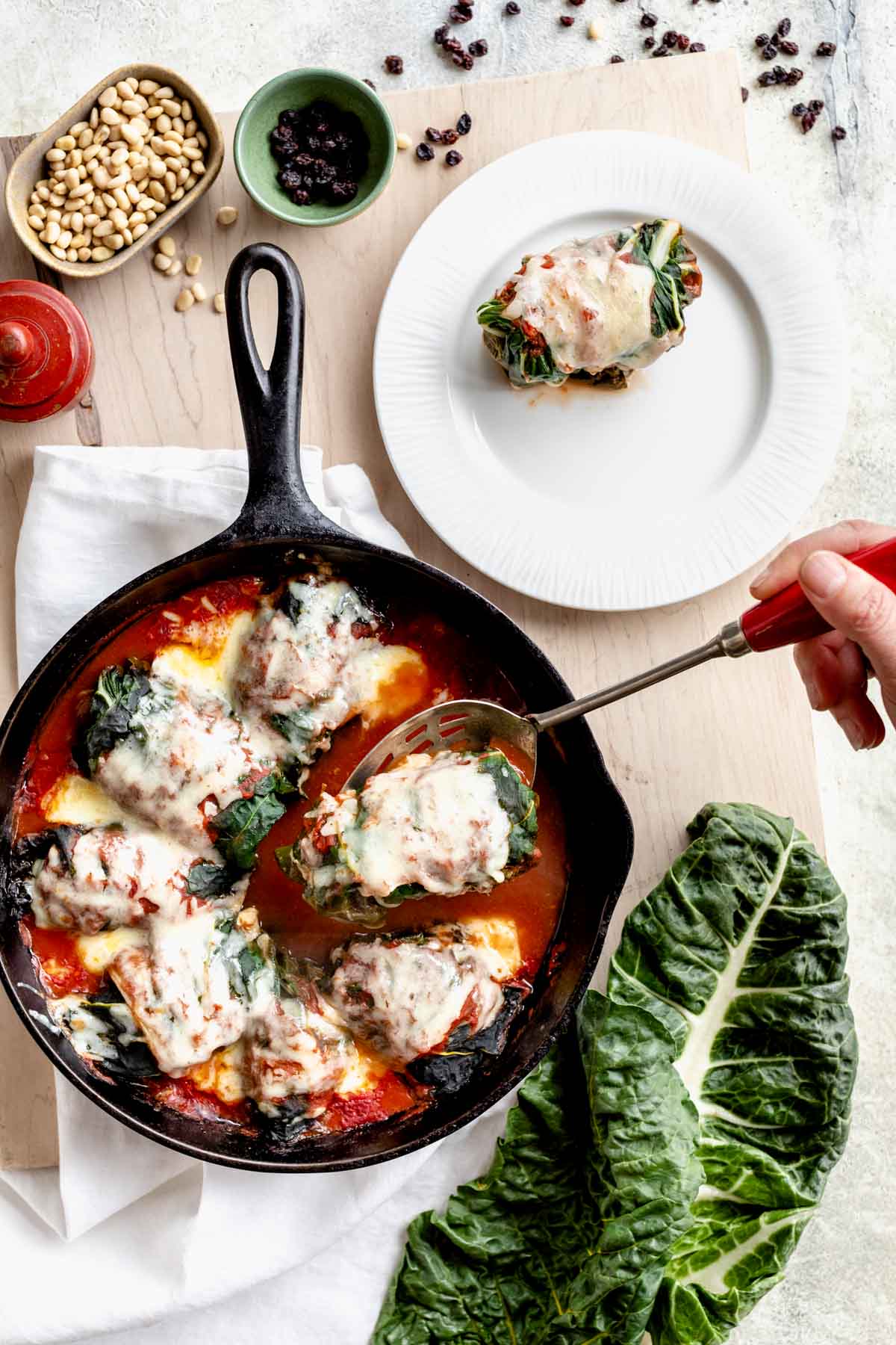 swiss chard rolls in a skillet, one is on a plate and another is being lifted up on a slotted spoon