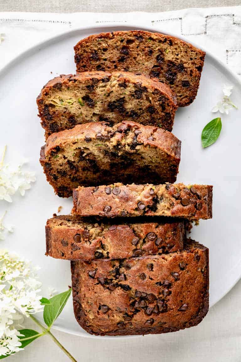 An overhead view of a loaf of zucchini chocolate chip bread sliced on a white platter with a hydrangea blossom