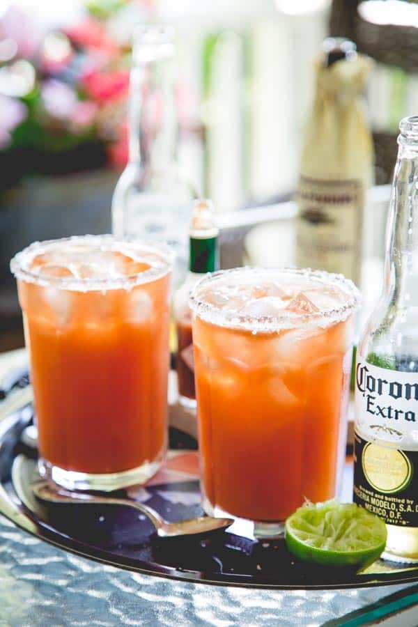 Two glasses of Michelada's on a black tray next to hot sauce, lime wedges and beer bottles