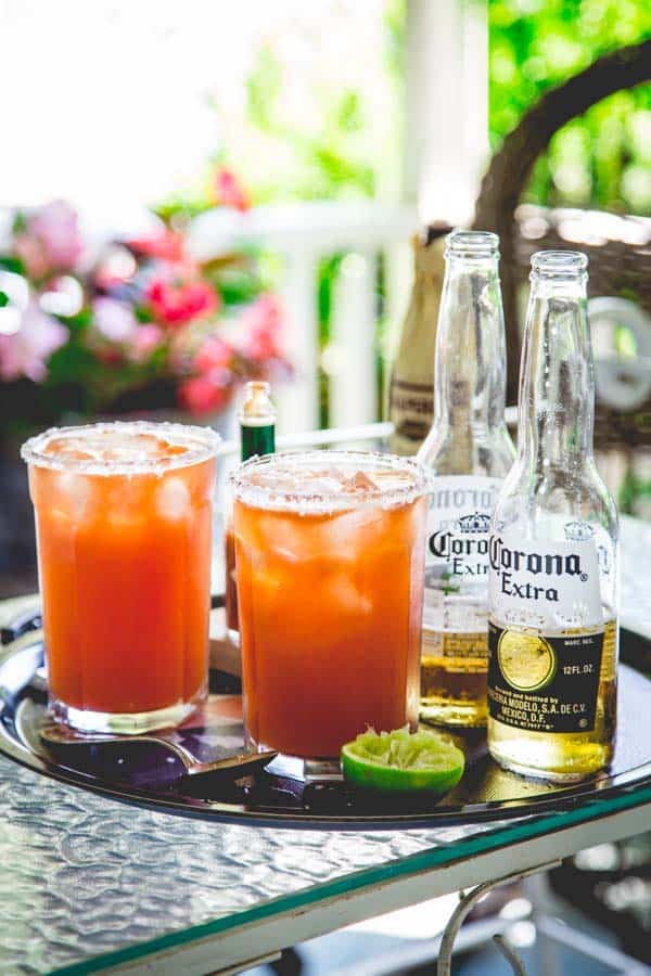Two glasses of Michelada's on a black tray next to hot sauce, lime wedges, spoon and beer bottles