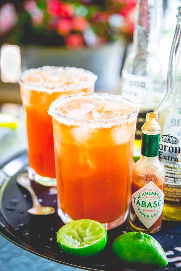 Two glasses of Michelada's on a black tray next to hot sauce, lime wedges and beer bottles