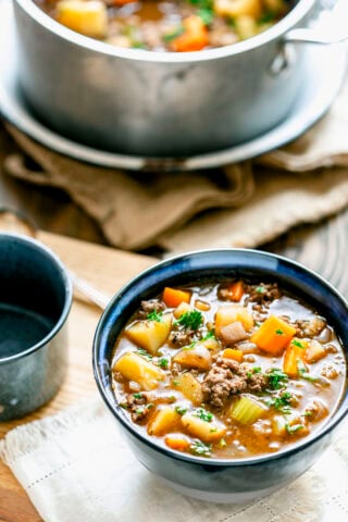 Delicious beef stew made with ground hamburger meat in a blue bowl with the soup pot in the background