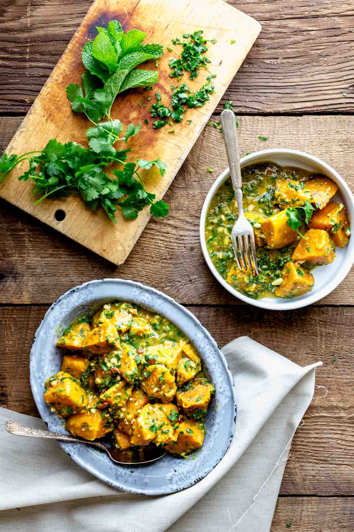 wooden table with a metal dish with sweet potato curry and a white bowl with a serving of the curry in it