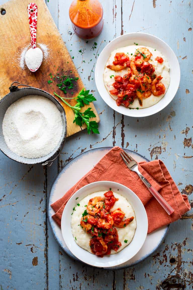 Two bowls of Spicy Shrimp and Cheese Grits on a blue surface next to a bottle of hot sauce and a bowl of grits on a cutting board with parsely