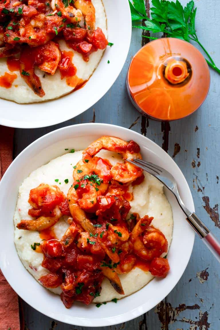Two bowls of Spicy Shrimp and Cheese Grits on a blue surface next to a bottle of hot sauce