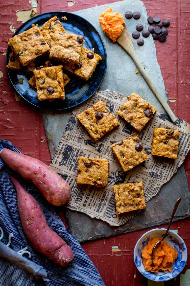 Sweet Potato Blondies on a blue plate and on newspaper on a slate board next to a small bowl of sweet potato puree, sweet potatoes, chocolate chips, a wooden spoon with sweet potato puree and a blue linen