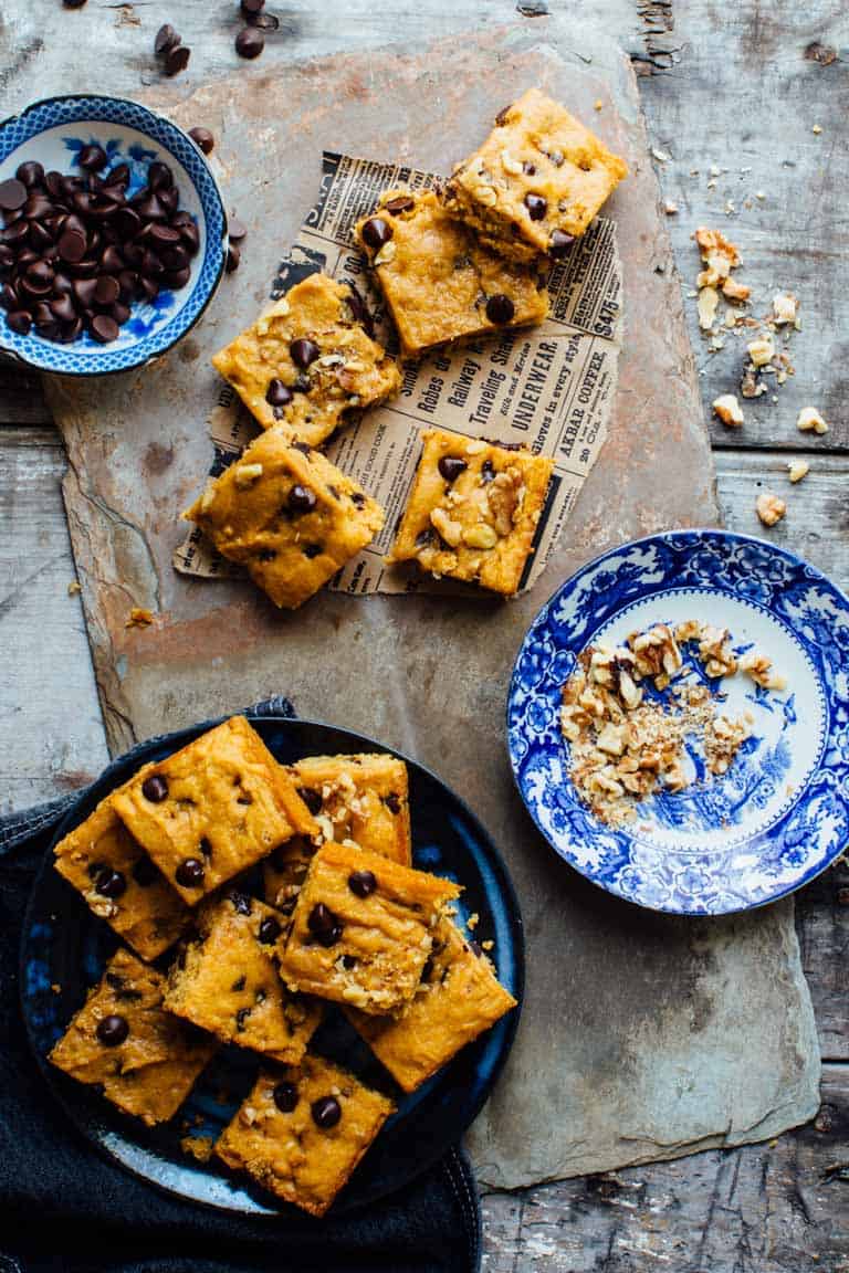 Sweet Potato Blondies on a blue plate and on newspaper on a slate board next to a small bowl of chocolate chips and a small bowl of walnuts