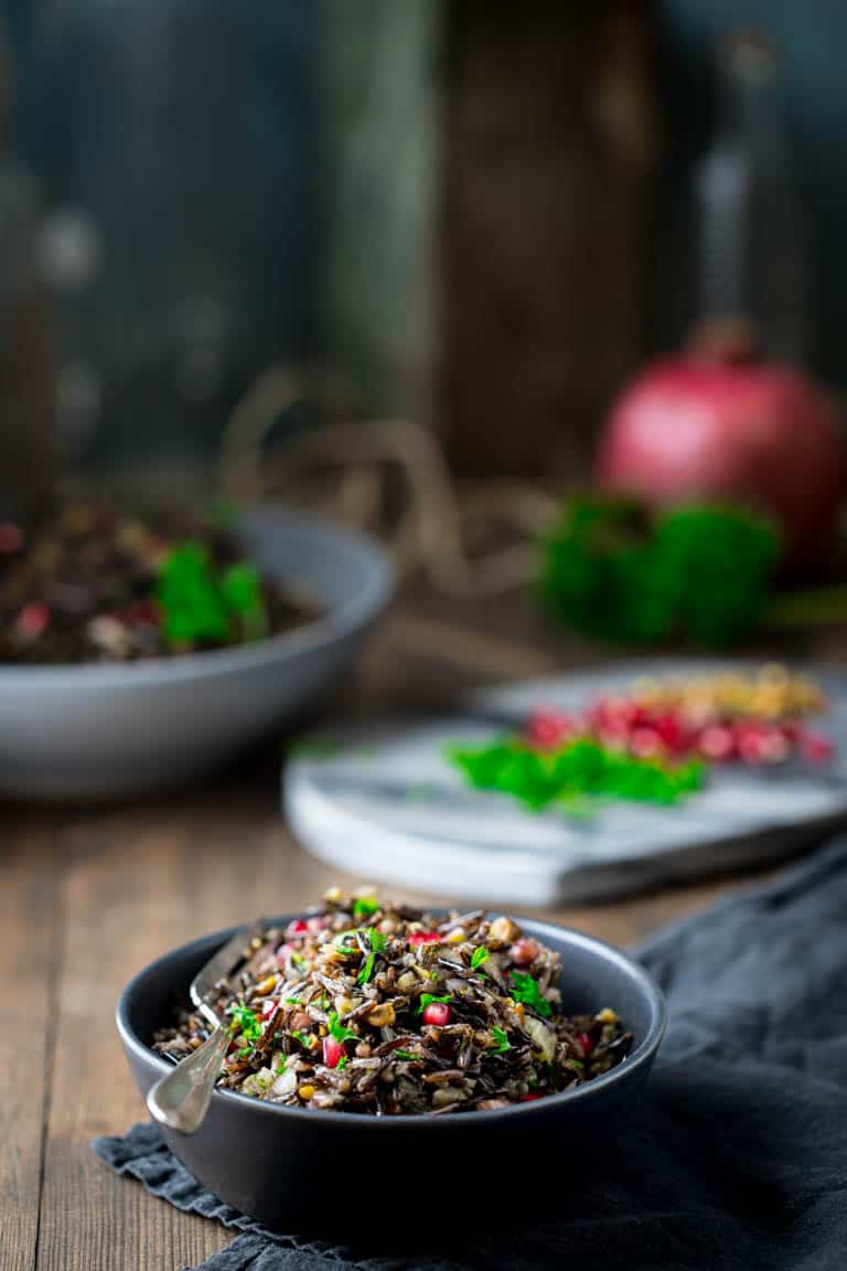 A serving bowl with wild rice salad with chopped parley, pistachios, and pomegranate seeds behind.