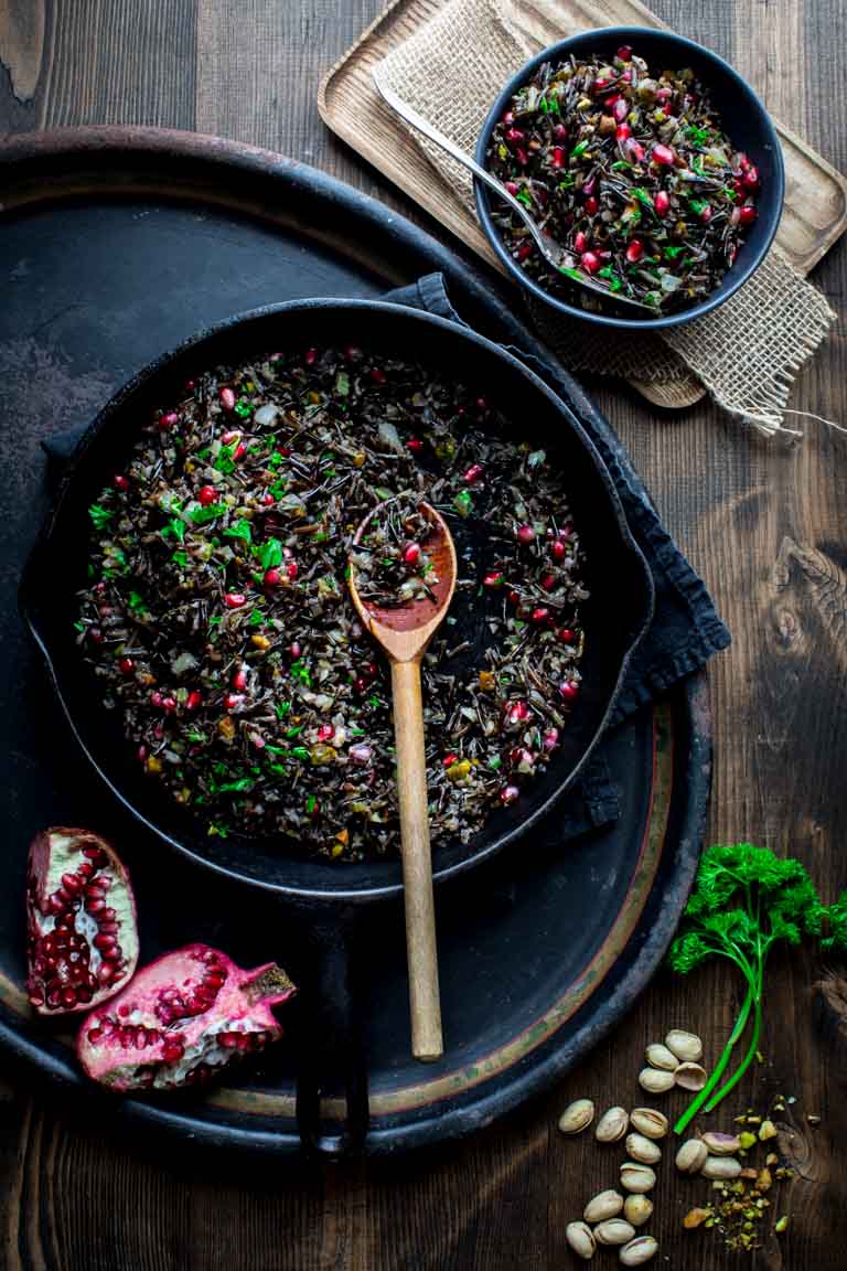 A full skillet of wild rice pilaf with pomegranates and parsley, with a serving in a bowl alongside.