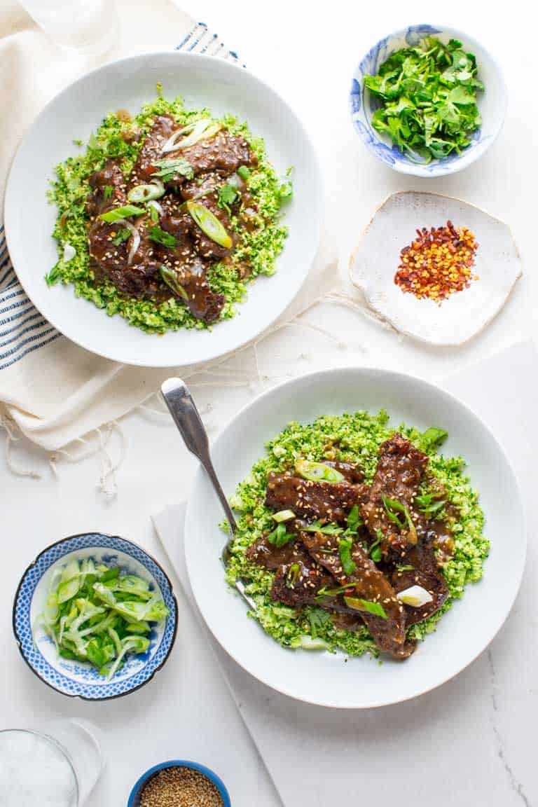 grain free mongolian beef and broccoli bowls beside smaller bowls of cilantro, red pepper flakes, and green onions for garnishing