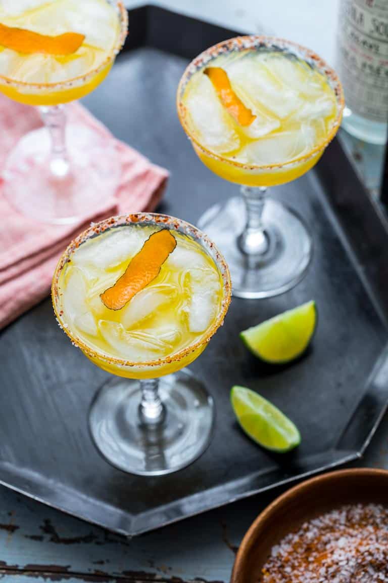 Over head photo of three Smoky Mescal Margaritas in coup glasses on a tray with a red linen and lime slices and small bowl of spiced salt
