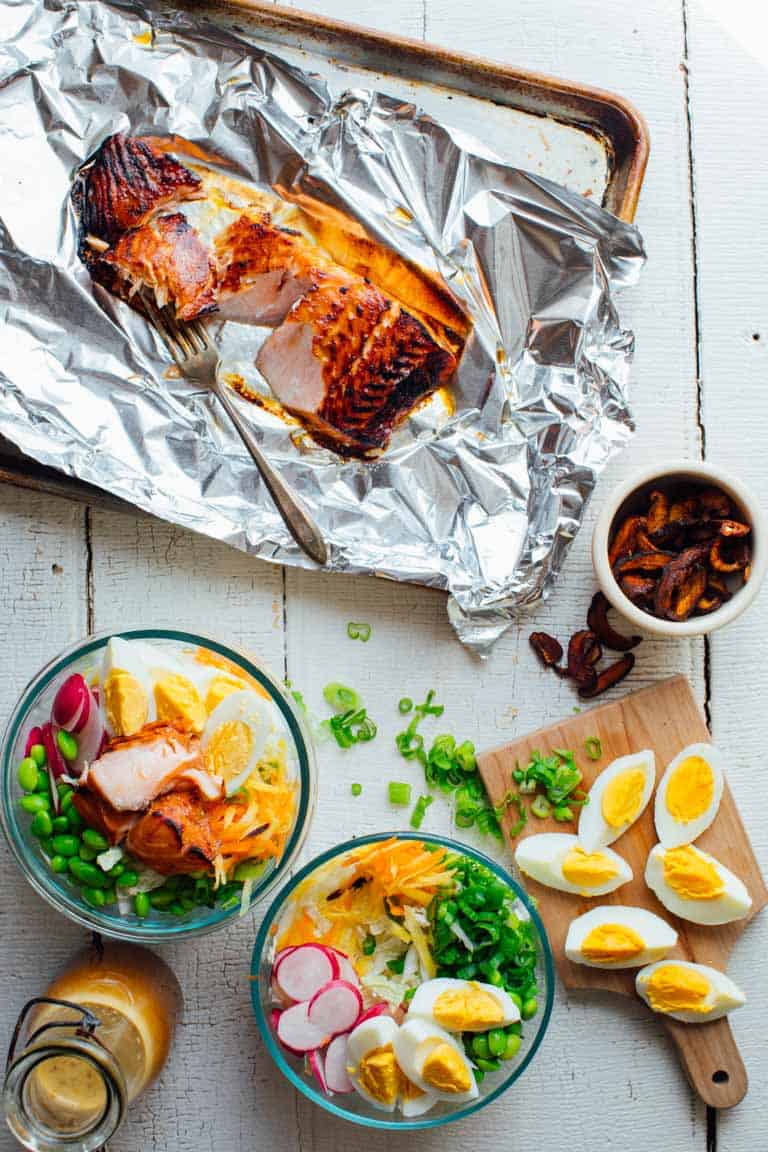 overhead shot of maple and wasabi glazed salmon on a baking sheet beside two bowls of meal prep power salad, hard boiled eggs, and shiitake mushrooms
