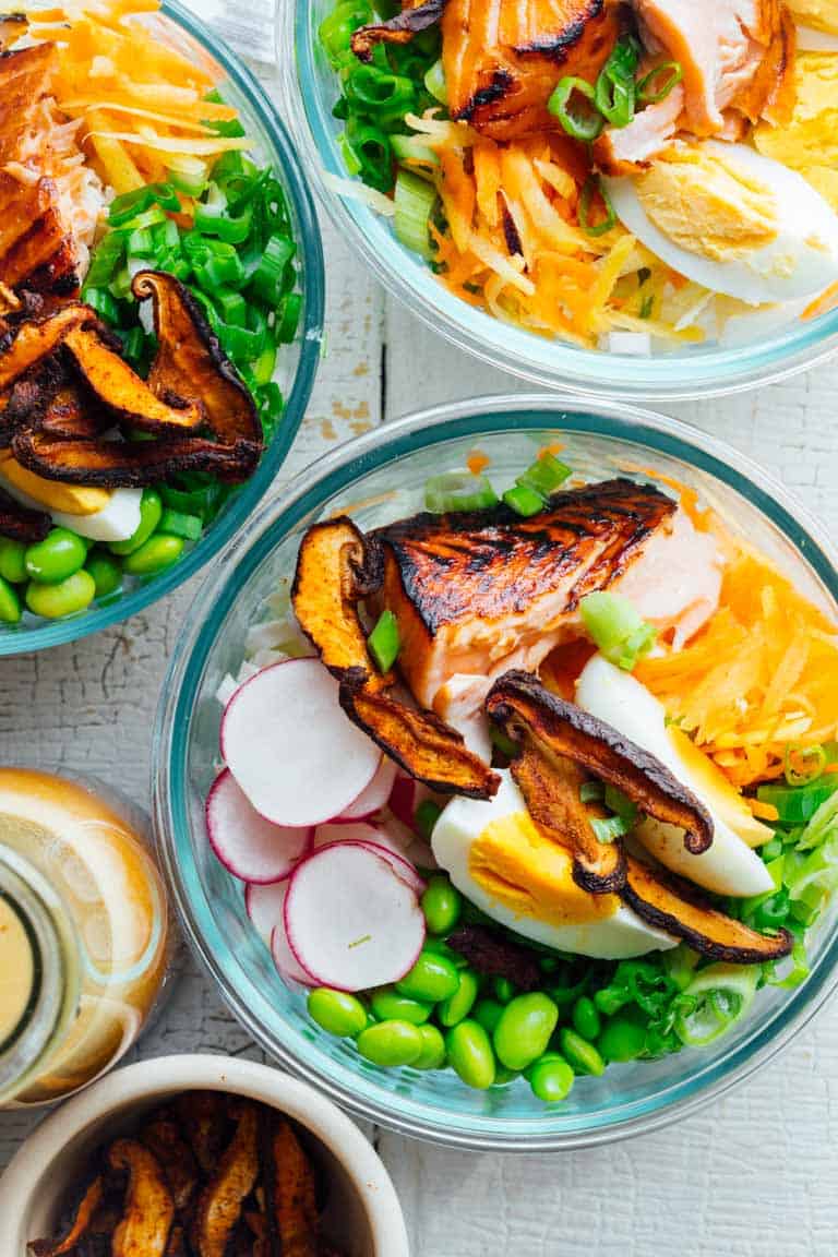A few bowls of meal prep salmon salads with wasabi glaze, hard boiled eggs, edamame, radishes, and shiitake mushroom bacon