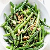 Green beans with balsamic and walnuts in a white serving bowl next to a small bowl of walnuts and a head of garlic
