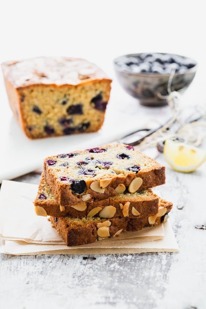 A stack of slices of blueberry bread next to a loaf and a bowl of blueberries
