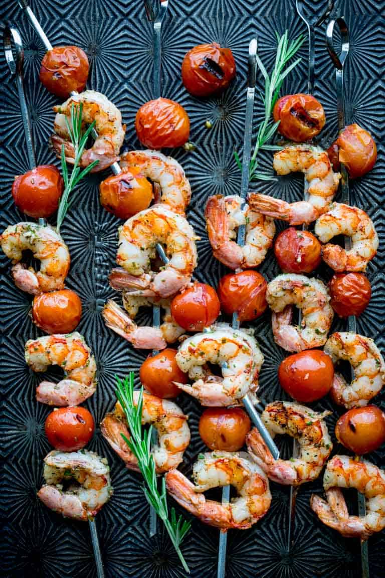 a black tray of shrimp kebabs with cherry tomatoes and rosemary