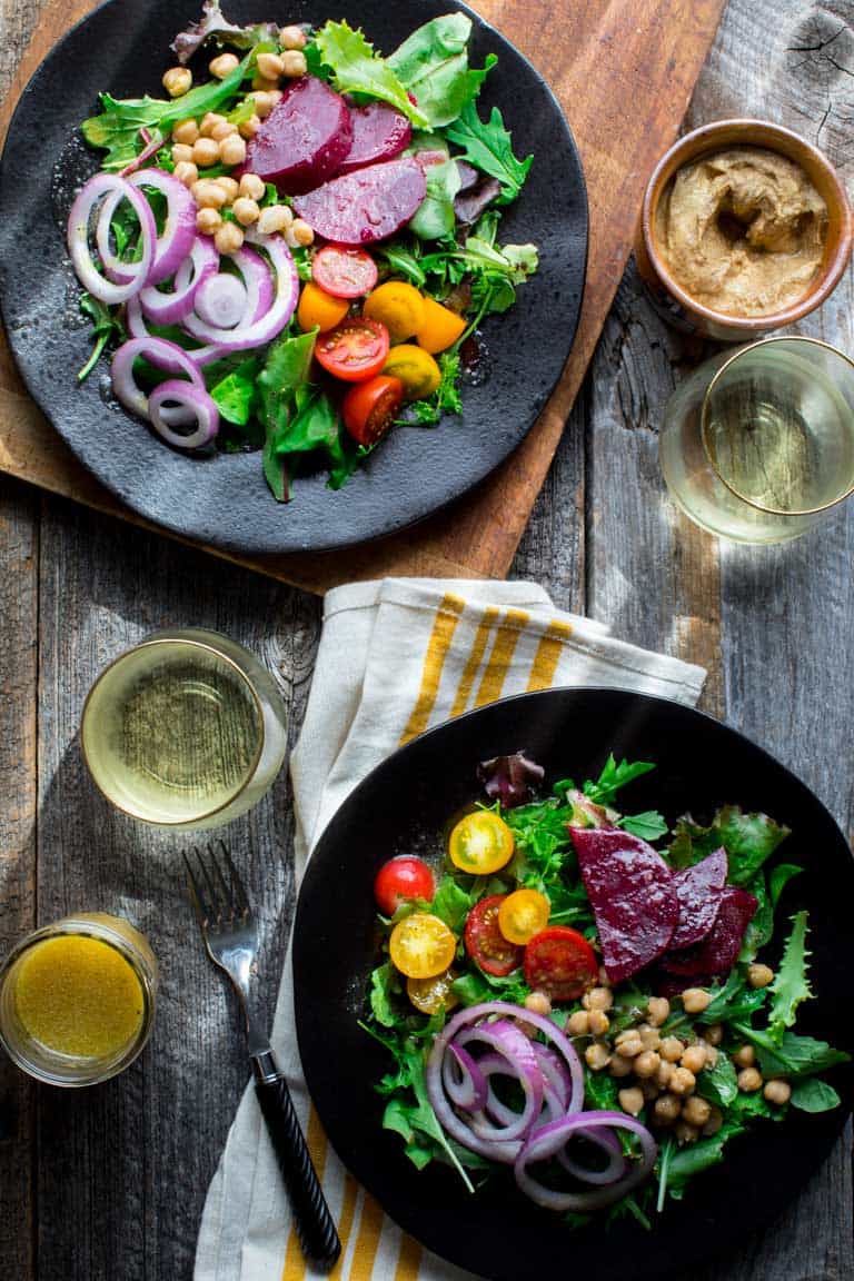 Two colorful salads with two glasses of white wine, a crock of german mustard and a jar of mustard salad dressing on a gray farm table