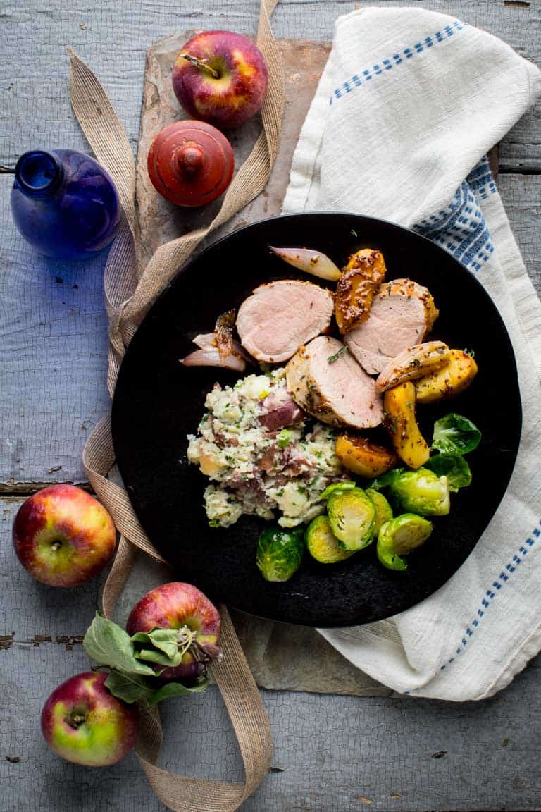 Easy Roasted Pork Tenderloin and Apple Skillet with Maple Mustard sauce and shallots. A grain-free and gluten-free one pan dish for entertaining or a weeknight! #porktenderloin #easy #onepan #entertaining a blue-gray painted table with a slate board. On top is a black plate with pork tenderloin, brussels sprouts and herb mashed potatoes